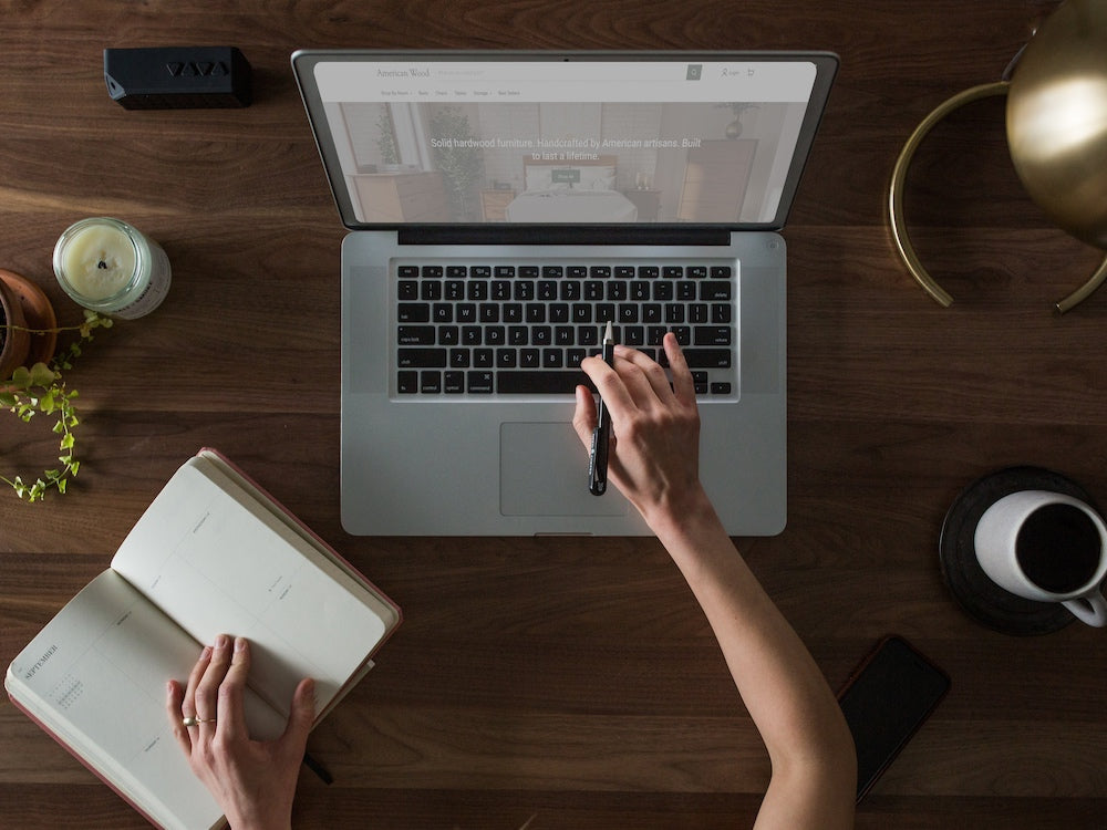 Top down view of a woman using a laptop on a wood table with an open notebook and coffee.